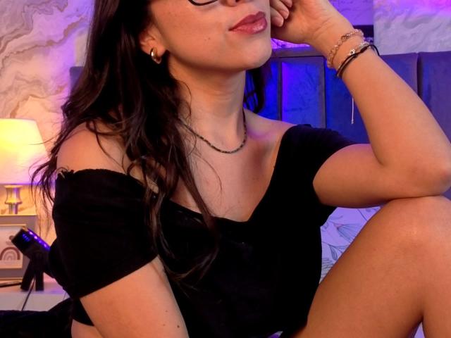 Mia Reed, avec ses cheveux foncés, ses lunettes et son haut noir à une épaule, assise avec une main sur son visage, montrant ses jolies fesses, dans une pièce faiblement éclairée..
