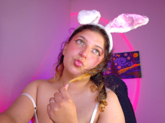 AmaliaRosse with curly brown hair, white bunny ears, pouty pink lips, green eyes, and a finger near her chin, wearing a white strap top, in a pink-lit room.