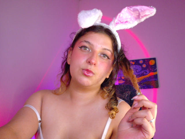 AmaliaRosse with curly brown hair, wearing white fluffy bunny ears, holding a black fan, pink lips puckered, light pink lipstick, light skin, pinkish-purple background, pink lighting.