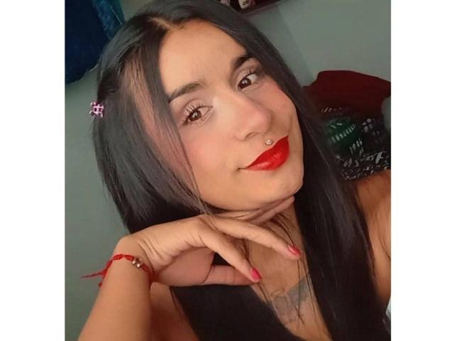 AmberLuxefg with long black hair, red lipstick, nose ring, and red nail polish, smiling slightly, holding her chin, wearing a red bracelet, looking cute and sexy.