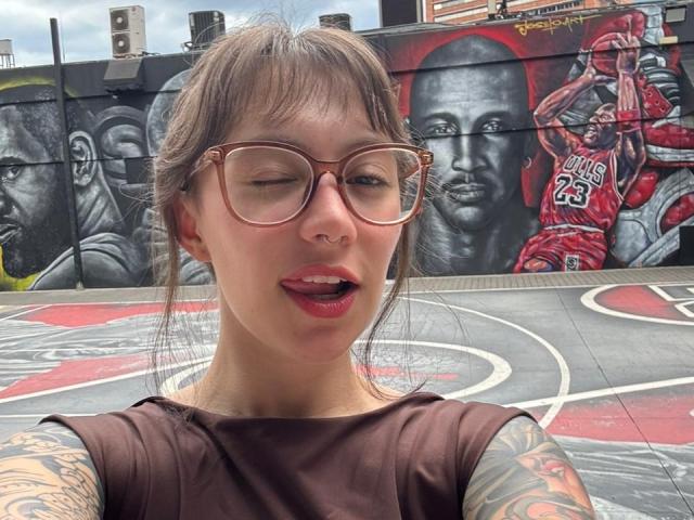 IsabellaArias has light skin, brown hair in a messy bun, and wears large, round, brown glasses. She has a nose ring and tattoos on her arms. Her tongue is out, and she looks slightly winking. She wears a brown sleeveless top. The background features a graffiti mural with black and red NBA jerseys and portraits of basketball players. The mural is detailed and vibrant, with a basketball court pattern. The image is a photograph.