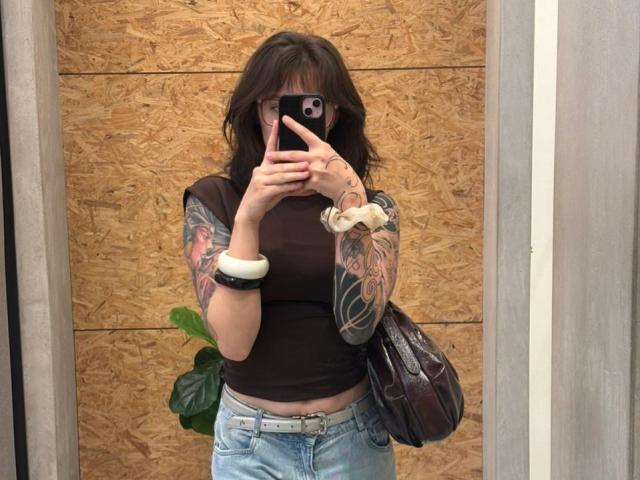 IsabellaArias stands in a mirror selfie. She has wavy, dark brown hair, and wears glasses. She's holding a black smartphone, partially covering her face. She wears a black, short-sleeve, crop top, revealing a white scrunchie on her right wrist. Her tattoos, colorful and detailed, cover both arms. She carries a black, patterned bag on her left shoulder. She's wearing light blue, slightly loose, jeans. The background is a wooden, textured wall. Her right hand is raised, with fingers pointing slightly upwards.