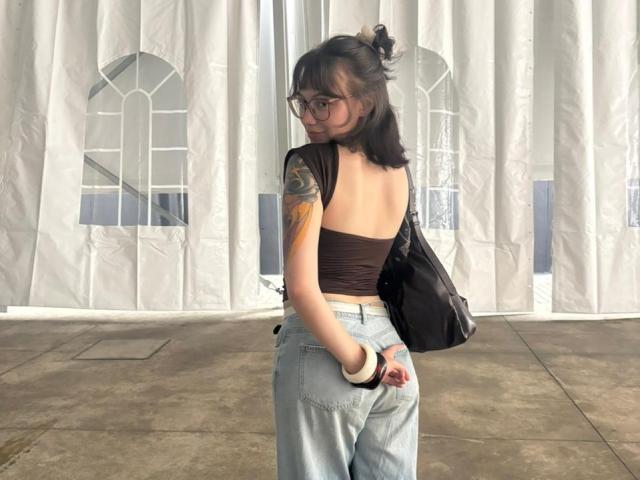 IsabellaArias with glasses, brown tube top, tattoo on left arm, light blue jeans, black bag, looking back, showing her ass, standing in front of white curtains.