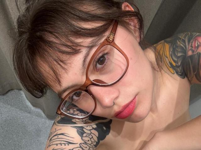 Close-up of IsabellaArias with short brown hair and glasses. She has a fair skin tone, pink lipstick, and a slight, confident smile. Her right shoulder and upper arm are covered in colorful, detailed tattoos. She's looking slightly upward, with her head tilted to the left. Her glasses are brown with a gold accent on the top right. The background is blurry and indistinct, with a gray curtain on the left and a gray carpet on the bottom. IsabellaArias' tattoos include a large floral design on her shoulder and a smaller tattoo on her arm. The image is focused on her face and upper torso.