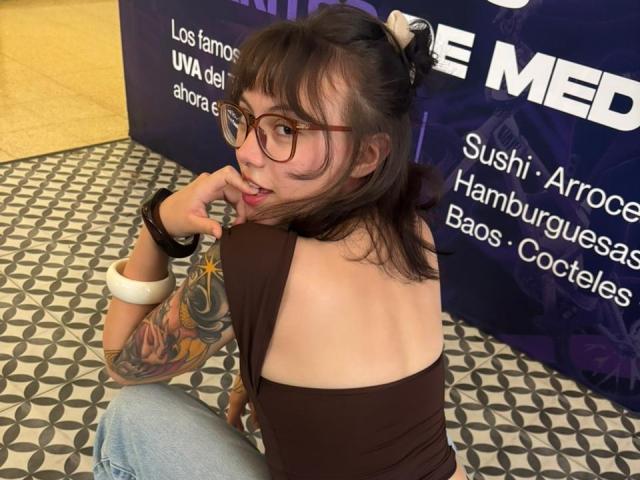 IsabellaArias with glasses, brown top, and tattoos on her right arm, looking over her shoulder with a finger near her mouth, sitting on a patterned floor.