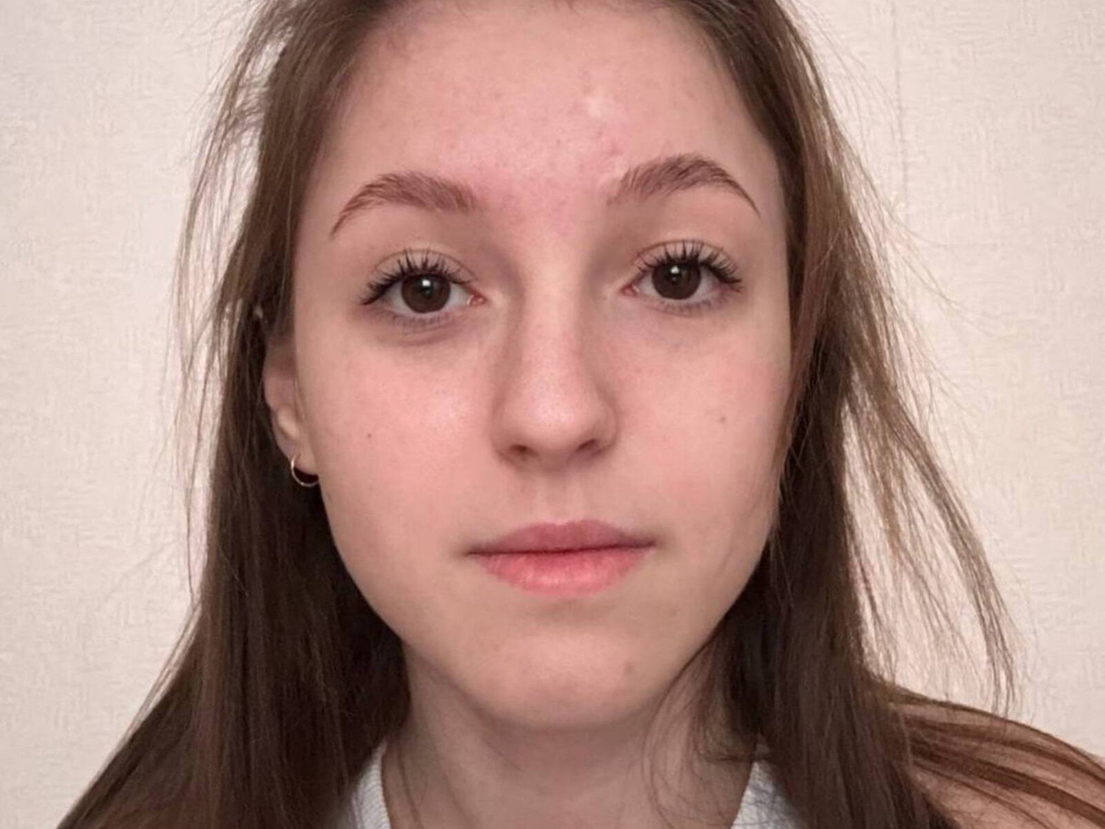 Close-up photograph of LucretiaEgnor with a neutral expression. She has light skin, brown eyes, and long brown hair slightly messy. Her eyebrows are thin and natural. She wears small hoop earrings and a white shirt. Her lips are slightly parted, and her eyelashes are long and dark. The texture of her skin is slightly bumpy with a few small blemishes. The background is a plain, textured beige wall. The image is well-lit, showing clear details of her facial features.