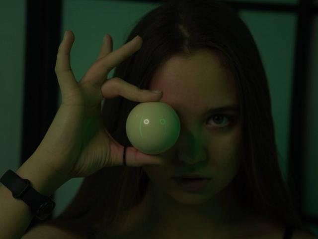 NickieZadd holds a green, glossy ball in front of her right eye with her right hand. Her dark brown hair is straight and falls over her shoulders. She wears a black wristwatch on her right wrist. The light is greenish, casting a shadow over her face. Her eyes are focused and slightly squinted. The ball has a subtle reflection, indicating its smooth surface. The image is dark and moody, emphasizing NickieZadd's intense expression.
