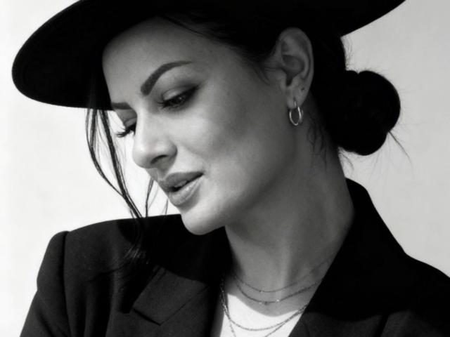 Black-and-white photo of VixenSelena with a hat, earrings, and a jacket, looking down with slightly parted lips, showing a delicate necklace. Her tits are partially visible.