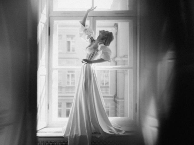 Black-and-white photo of EolantaMarina standing in a window, arms raised, wearing a flowing dress, her tits slightly visible, back to the light, soft shadows, elegant pose.