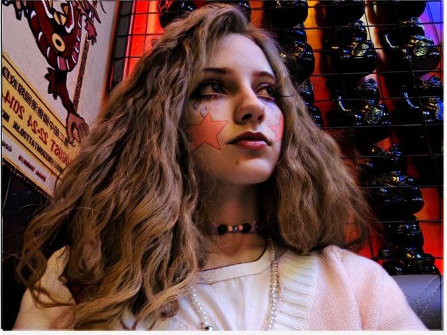 CuteeFoxy has wavy, medium-brown hair and a star painted on her right cheek. She wears a white shirt, a beaded choker, and a pearl necklace. Her expression is slightly raised, looking to the right. The background features a grid of black, shiny, horn-like objects on a red wall. The left has a poster with text and a red owl illustration. The image has a blue light reflection. Slightly blurry.