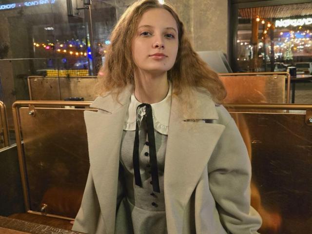 CuteeFoxy has wavy, shoulder-length brown hair and a small cut on her right cheek. She wears a light gray coat over a gray dress with black ribbon details and a white collar. Her expression is neutral, and she sits in a dimly lit, metallic booth. Warm, out-of-focus city lights are visible in the background. The composition uses warm, muted colors and soft light, highlighting her face and coat. The texture of the coat and dress contrasts with the metallic booth.