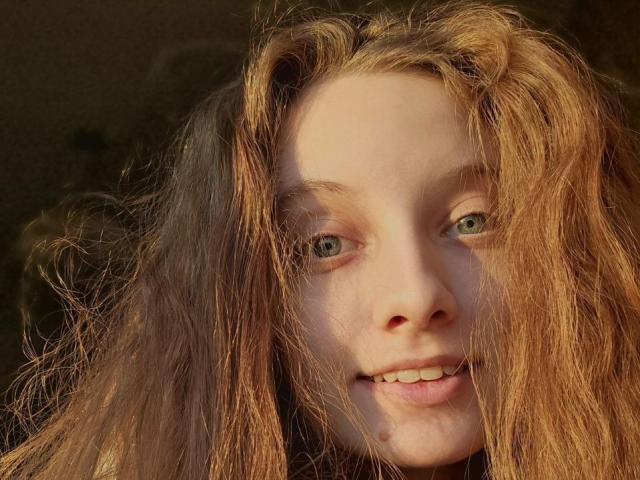 Close-up of CuteeFoxy with wavy, light brown hair partially covering her face. She has pale skin, green eyes, and a slight smile with visible teeth. Her hair is unkempt, with strands falling over her forehead and cheeks. Soft light illuminates her face, highlighting the natural texture of her hair and skin. The background is dark and out of focus, emphasizing CuteeFoxy's facial features and expression. The image has a candid, intimate feel.