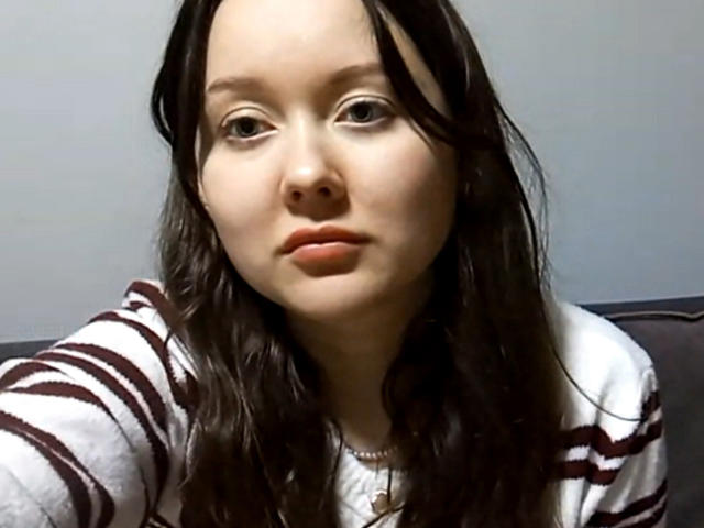 EvelineZabel has a pale face, brown wavy hair, and wears a striped white and brown shirt, with a neutral expression and slightly parted orange lips.