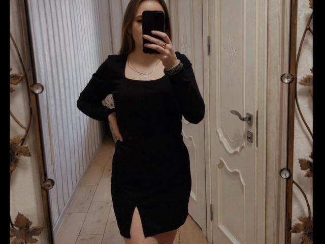 Lizza69 in a black dress with long sleeves and a thigh-high slit, taking a mirror selfie with a phone. Boobs visible under the dress, hand on hip.