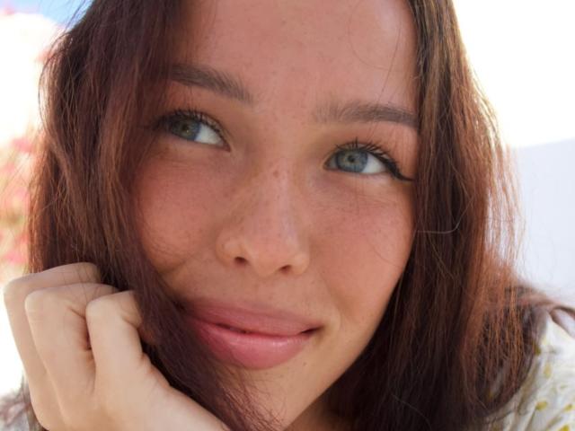 Close-up of JaneWow's face. She has light brown, slightly wavy hair, blue-green eyes, and fair skin with freckles. Her right hand is resting under her chin, with her fingers slightly curled. She's looking to the left with a subtle, relaxed smile. The image has natural light, and her lips are slightly parted. The background is blurred, emphasizing JaneWow's facial features. The texture of her hair is soft and slightly messy. She's wearing a white shirt with a small yellow pattern.
