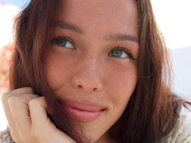 Close-up of JaneWow's face. She has light brown, slightly wavy hair that falls around her face. Her blue-green eyes are looking to the side. She has a small, gentle smile with slightly parted pink lips. Her right hand is resting on the side of her face. She has light freckles on her cheeks and nose. Her eyebrows are thin and slightly arched. The image is brightly lit, with natural light highlighting her features. The background is blurred, focusing attention on JaneWow's face. Her makeup is minimal, with a subtle eyeliner. The texture of her hair is slightly rough.