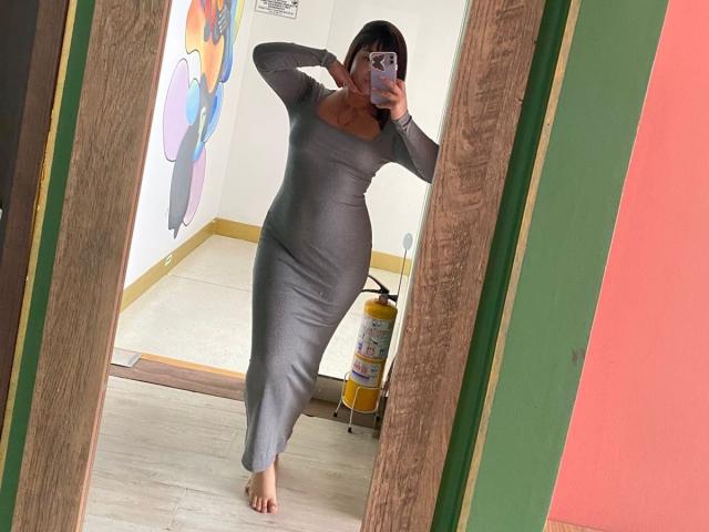 AnikaFleur stands in a mirror, taking a selfie. She's wearing a tight, long-sleeve, gray dress that hugs her curvy, hourglass figure. Her right hand is near her face, holding a phone with a purple case. She has dark hair in a bob cut. The wooden frame of the mirror is visible. The room has colorful abstract art on the left wall and a yellow fire extinguisher on the right. The floor is light wood, and she's barefoot. The mirror reflects a room with a green and pink wall.