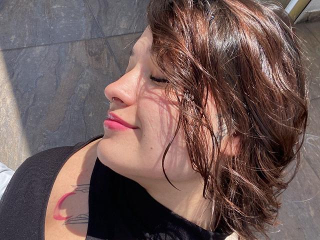 AnikaFleur's face, with pink lips and brown wavy hair, showing a tattoo on her right shoulder, eyes closed, smiling, sunlit, black top.