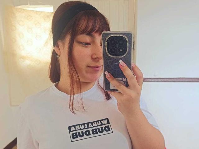 NatalieDyer with brown hair, white shirt, black headband, holding phone, taking selfie, pouty lips, small nose, medium tits, light skin, pink nails, white wall in background.
