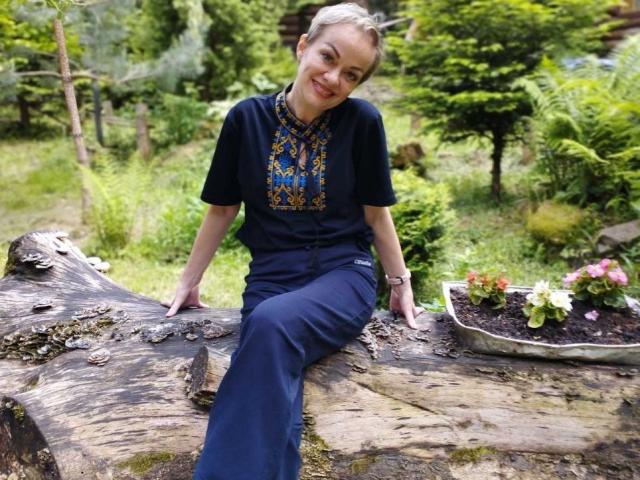 NikaMayu sits on a rough, moss-covered log with her legs extended, smiling at the camera. She wears a navy blue shirt with a colorful, embroidered pattern and matching pants. Her blonde hair is pulled back. A rectangular planter with pink and white flowers is to her right. The log has a textured, weathered surface with visible grain patterns. NikaMayu's relaxed, confident pose contrasts with the log's rugged texture. The bright greenery in the background enhances the natural, outdoor setting.