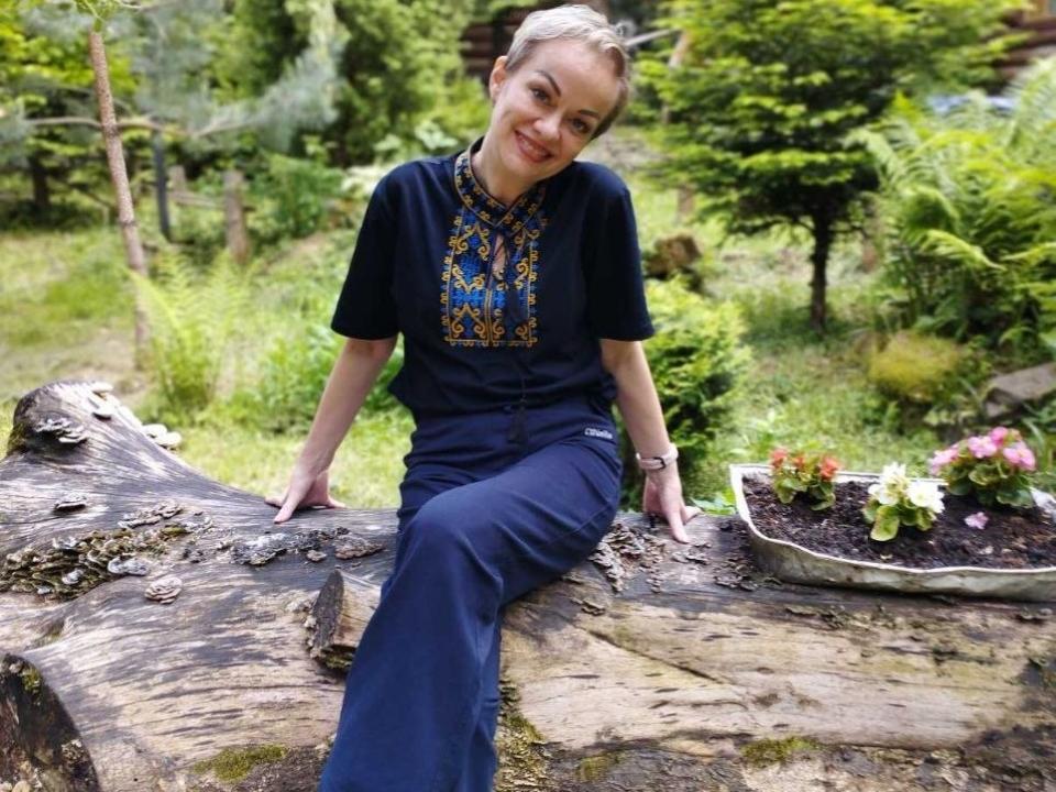 NikaMayu sits on a rough, moss-covered log with her legs extended, smiling at the camera. She wears a navy blue shirt with a colorful, embroidered pattern and matching pants. Her blonde hair is pulled back. A rectangular planter with pink and white flowers is to her right. The log has a textured, weathered surface with visible grain patterns. NikaMayu's relaxed, confident pose contrasts with the log's rugged texture. The bright greenery in the background enhances the natural, outdoor setting.