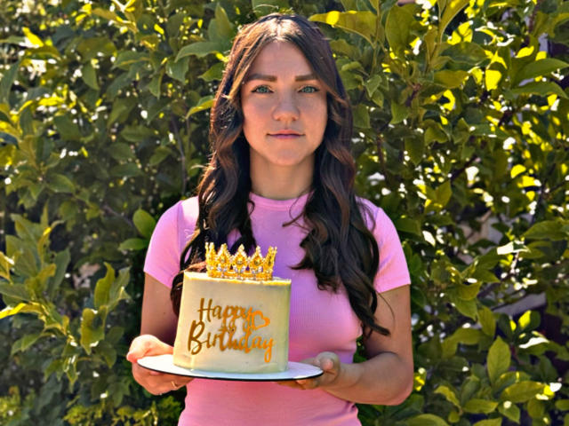 CammieRosalie with wavy brown hair, pink shirt, holding yellow birthday cake with gold crown, "Happy Birthday" in orange, standing in front of green leafy background.