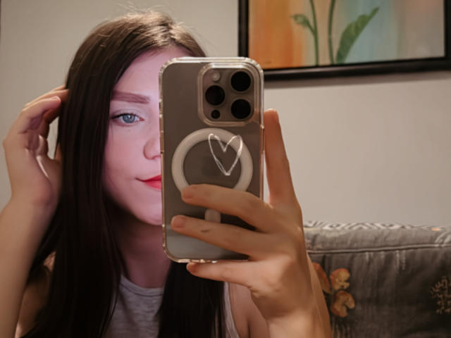 CammieRosalie takes a selfie with an iPhone in a well-lit room. She has long, straight brown hair, fair skin, and blue eyes. She's wearing a grey shirt. Her right hand holds the phone, which has a grey case with a white heart ring. The background includes a floral-patterned couch and a framed painting with green and orange colors. The composition is centered on her face and phone.