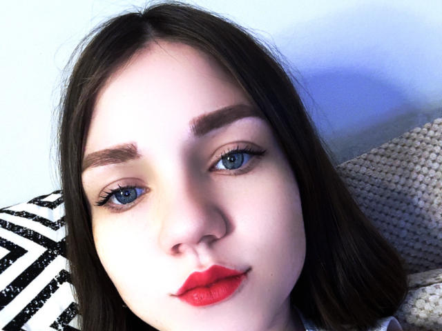 Close-up of CammieRosalie's face. She has fair skin, blue eyes, and dark brown eyebrows. Her long, straight brown hair falls over her shoulders. She wears bright red lipstick and has long, dark eyelashes. She's looking directly at the camera with a slight smile. The background includes a black and white striped pattern on the left and a textured beige fabric on the right. The lighting is even, highlighting her facial features.