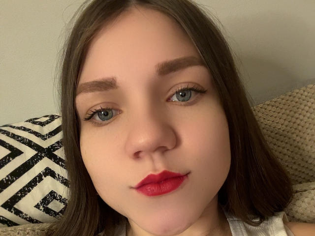 Close-up of CammieRosalie, a young woman with fair skin and straight brown hair. She has blue eyes, thick eyebrows, and wears red lipstick. Her eyelashes are long and dark. She is looking directly at the camera with a slight, confident smile. She has a small, subtle nose and a soft, youthful face. The background includes a beige textured surface and a black and white patterned pillow. The image has a slightly warm, natural light.