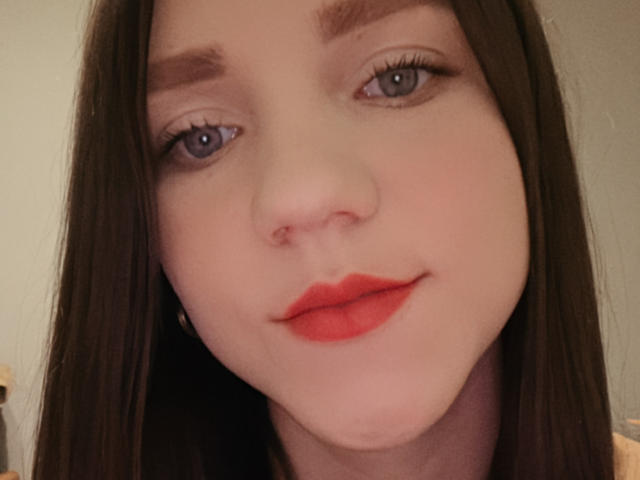 CammieRosalie's face, showing pale skin, blue eyes, dark brown eyebrows, long brown hair, red lipstick, and a subtle smile. Her nose has a small pinkish nose ring.