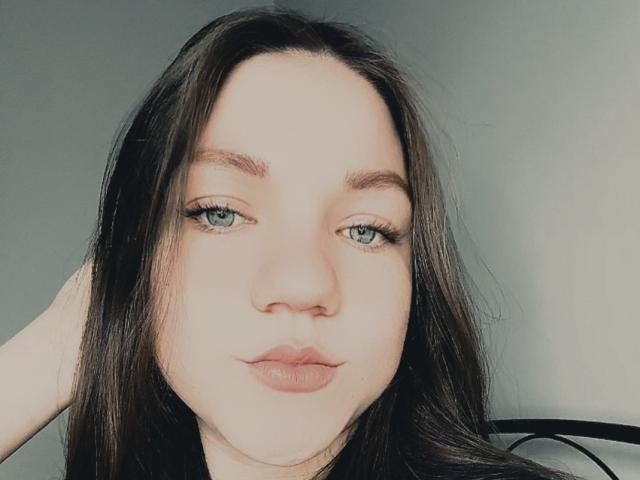 Close-up portrait of CammieRosalie with fair skin and long, straight black hair. Her green eyes are slightly open, and she has full, pink lips. She has minimal makeup, with subtle eyeliner and mascara. Her eyebrows are dark and well-defined. She's looking directly at the camera with a neutral expression. The light is soft, creating a slight shadow on her right cheek. The background is a plain, light gray. Her right hand is partially visible, resting behind her head. The overall composition is centered and balanced.