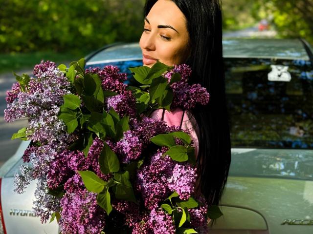 Xioni with long black hair, holding a big bouquet of purple lilacs, standing in front of a white car, looking down with a slight smile, sunny day.