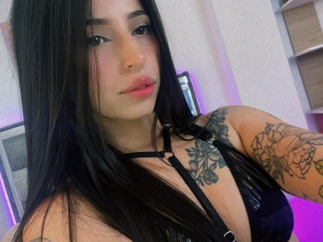 AllissonX, con cabello negro, aro en la nariz y tatuajes, vestida con un top de cuero negro estilo halter que deja ver sus senos, mirando a la cámara con labios rosados..