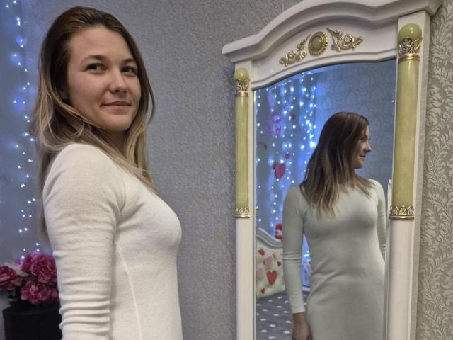 Anshante with blonde hair, white dress, and small tits standing beside a mirror reflecting her. She's smiling, looking right. Mirror has gold accents, blue fairy lights in background.