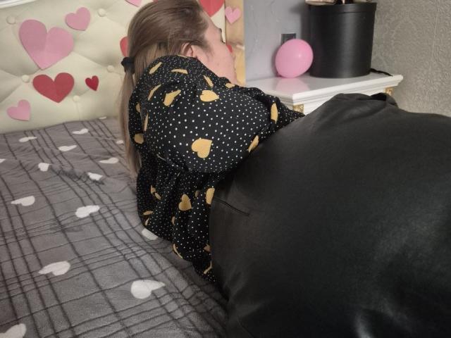 Anshante lies face down on a bed, wearing a black polka dot shirt with yellow hearts, her ass raised, black leather blanket covering her. Her blonde hair is visible.