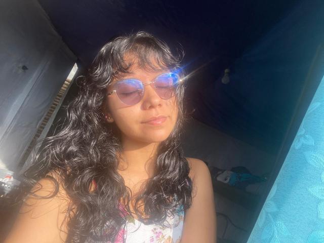 JuliaBennett, a young woman with long, wavy black hair, wears pink-tinted aviator sunglasses, basking in sunlight. She has a light brown skin tone, subtle makeup, and a calm expression. She's dressed in a white floral tank top. Sunlight creates a lens flare effect on her sunglasses. The background is dark and out of focus, with a blue curtain on the right. Her hair is slightly messy, and the sunlight enhances the texture.