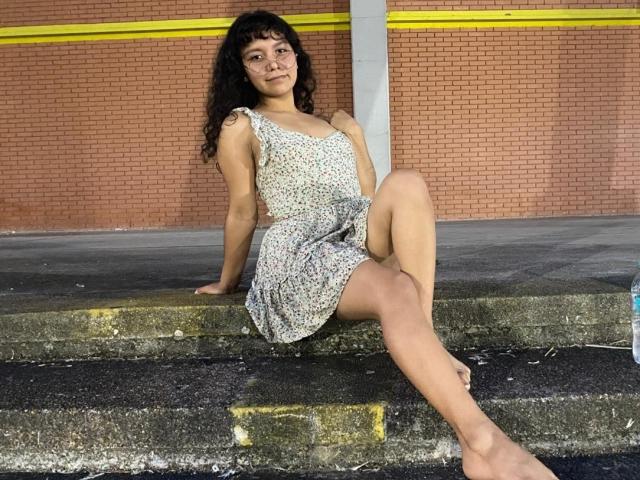 JuliaBennett sits on concrete steps with her right arm resting on her knee and her left hand on the step. She wears a floral sleeveless dress, exposing her bare feet. Her curly dark hair is loose, and she looks slightly to the left. Her skin is light brown, and she has small tattoos on her right shoulder. The background is a brick wall with yellow horizontal lines. The steps are textured with green and yellow stains. The image has a casual, relaxed vibe.