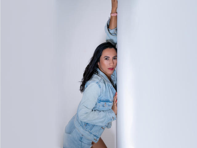 JadeFoster with black hair, wearing a light blue denim jacket and matching shorts, leaning against a white wall with one arm up, looking at the camera.