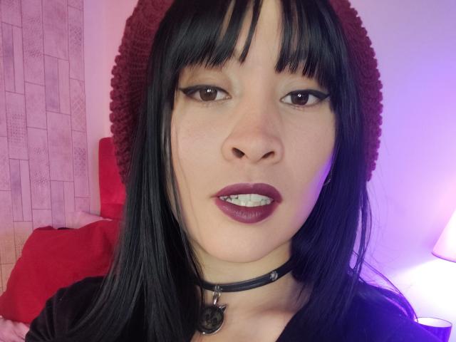 IssisDiosa has black hair, bangs, and a red beanie. She's wearing dark lipstick, a black choker with a pentagram, and a black shirt. Her teeth are slightly visible.
