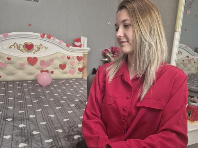 Anshante with blonde hair, wearing a red button-up shirt, sitting in a room with a heart-patterned bed and polka-dot gray blanket. Her tits are small, she's looking down.