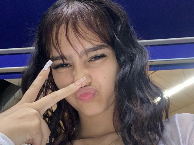 MelodyMansson with messy black hair, making a duck face, holding a finger to her nose. She's got long eyelashes and a white shirt. Her lips are pouty.