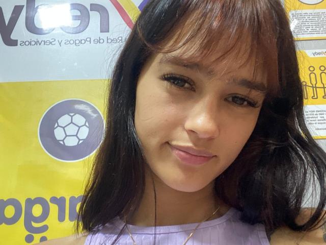 MelodyMansson has brown eyes, dark straight hair with bangs, light brown skin, and a slight smile. She's wearing a purple shirt, with a gold necklace visible.