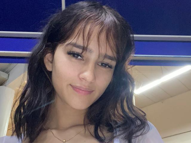 MelodyMansson has wavy brown hair, light skin, and a small nose. She's smiling with subtle makeup, wearing a white top, and a thin necklace. Her tits are not visible.