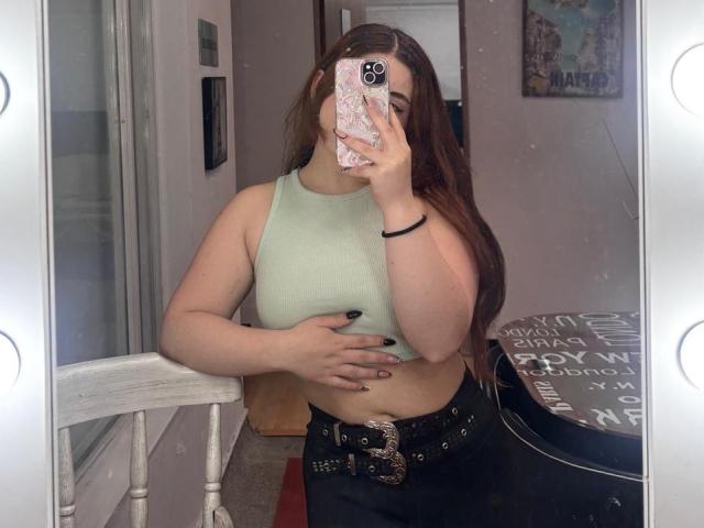 VicluX stands in front of a mirror, taking a selfie with a phone that has a pink, floral-patterned case. She has long brown hair and wears a light green sleeveless crop top. Her right hand holds the phone, covering part of her face, while her left hand rests on her stomach. She wears black pants with a studded belt. The mirror has light bulbs on the frame, reflecting her image. A wall with a small picture and a door are visible in the background.
