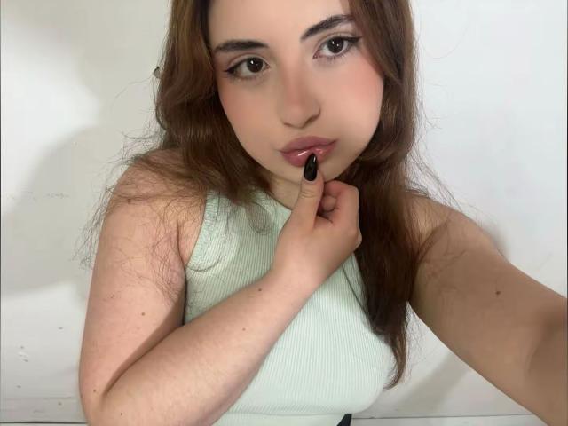 VicluX has long brown hair, slightly wavy, and light skin. She's looking directly at the camera with brown eyes. Her lips are slightly parted, and she's touching her lower lip with a black-painted fingernail. She's wearing a light green, sleeveless, ribbed top. The image is a close-up, focusing on her face and upper torso. Her expression is neutral. The background is plain and white.