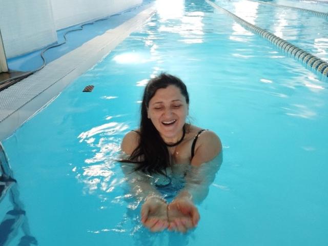 BlissSophia, a woman with long dark hair, is in a swimming pool with bright blue water. She is smiling with eyes closed, holding her orange-painted hands in front of her. She is wearing a black one-piece swimsuit and a black choker. The pool has a white tiled edge and a blue lane divider on the right. Light reflects off the water, creating small ripples. BlissSophia is centered in the image, with her face and upper body visible. The overall composition is bright and cheerful.