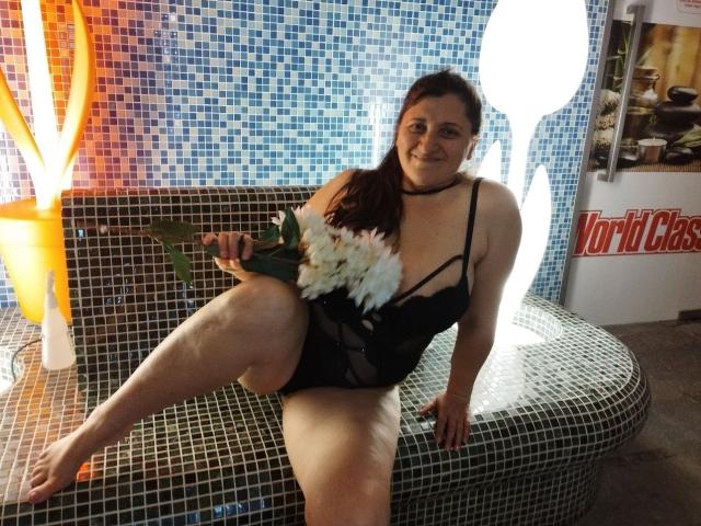 BlissSophia sits on a tiled bench, holding white flowers. She has pale skin, dark brown hair in a ponytail, and wears a black lace lingerie bodysuit with thin straps. Her right leg is raised, showing a thick, curvy thigh. The background has blue mosaic tiles, a glowing orange lamp, and a "Vodka Class" sign. The image is casual and slightly blurry. BlissSophia's expression is relaxed, with a subtle smile.