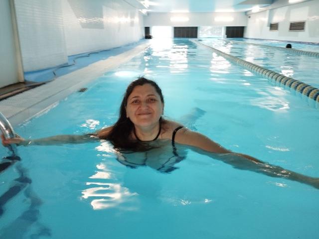 BlissSophia, a woman with long dark hair, is in a swimming pool, holding a metal ladder with both hands. She's wearing a black swim top, and her arms are stretched out. She's smiling at the camera. The pool water is bright blue, and a lane marker is visible to the right. The pool area is white-tiled with bright overhead lights. BlissSophia is centered in the frame.