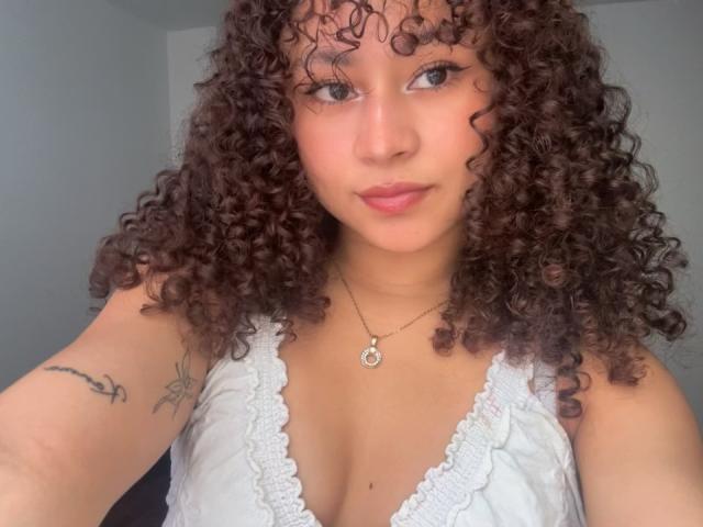 SofiaPassionne with curly brown hair, light brown skin, small tits, white lace top, tattoo on left arm, necklace, pink lips, and a slight smile in a selfie.