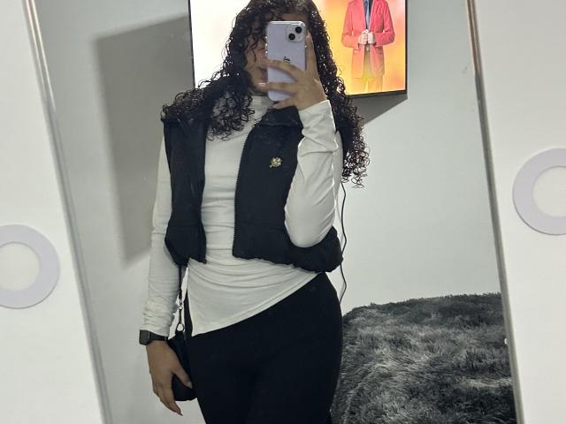SofiaPassionne with curly hair, wearing a black vest over a white long-sleeve shirt and black pants, taking a mirror selfie with her phone, showing her ass.
